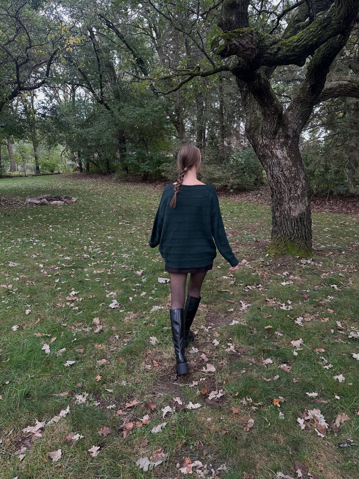 A person in a dark sweater, mini skirt, and tall black boots walking through a wooded area.