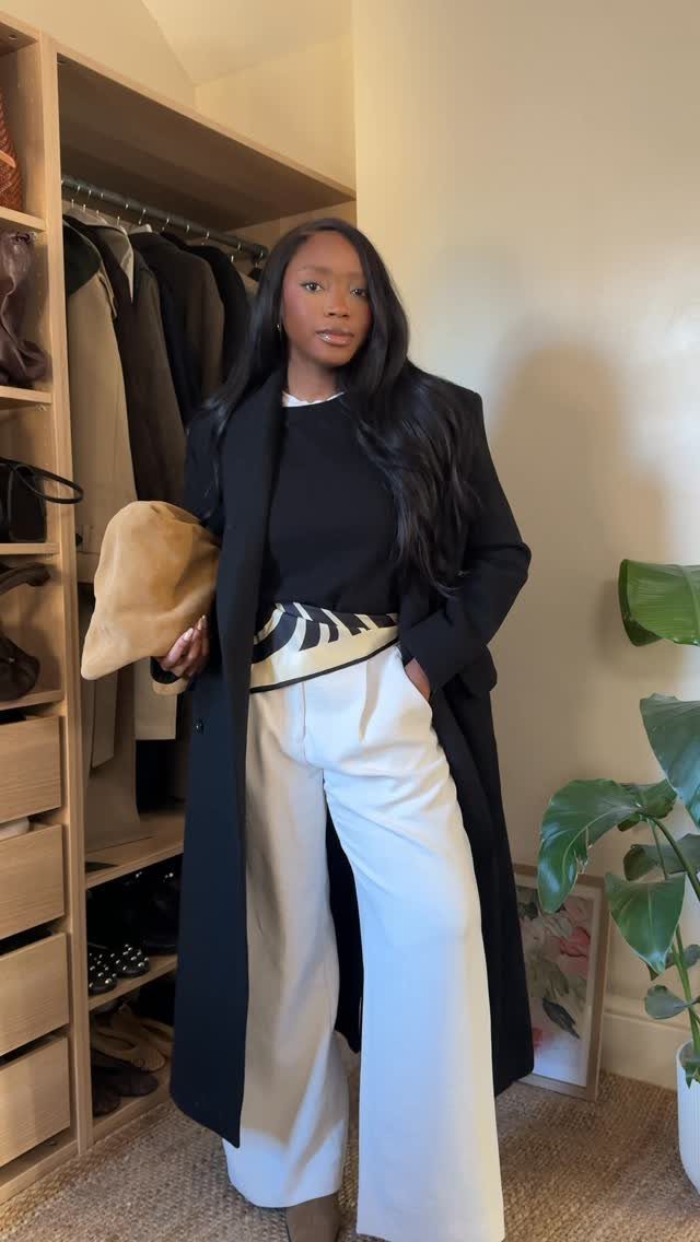 Woman wearing a black long coat, black top, and wide-leg white trousers in a closet.