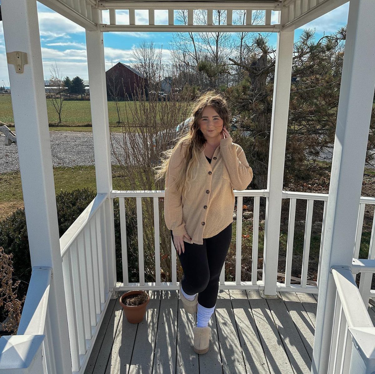 Woman in a tan checkered shirt and black leggings standing inside a white wooden gazebo.