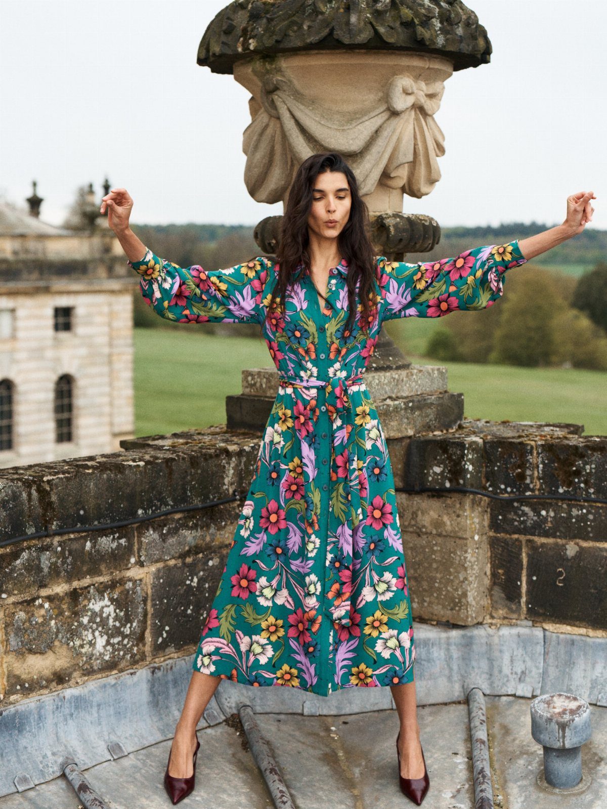 Woman in a teal floral maxi dress posing on a stone balcony overlooking a landscape.