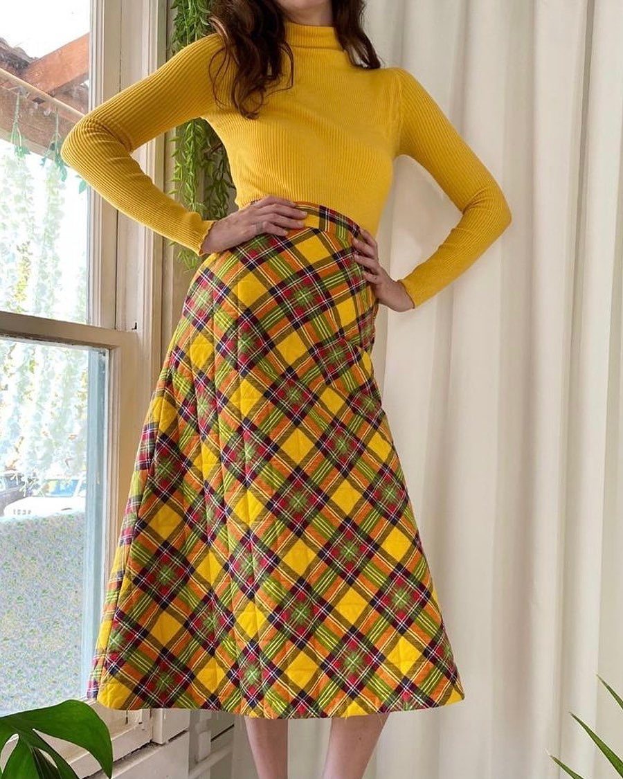 A person wearing a yellow turtleneck and a yellow plaid midi skirt standing by a window.