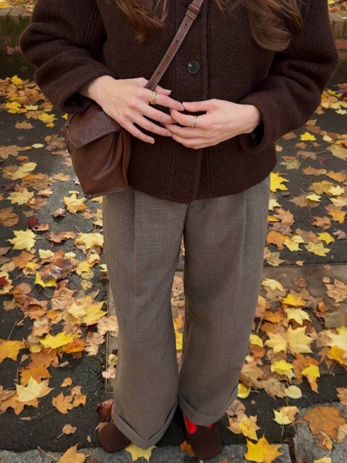 A person wearing a brown jacket and grey trousers standing on autumn leaves.