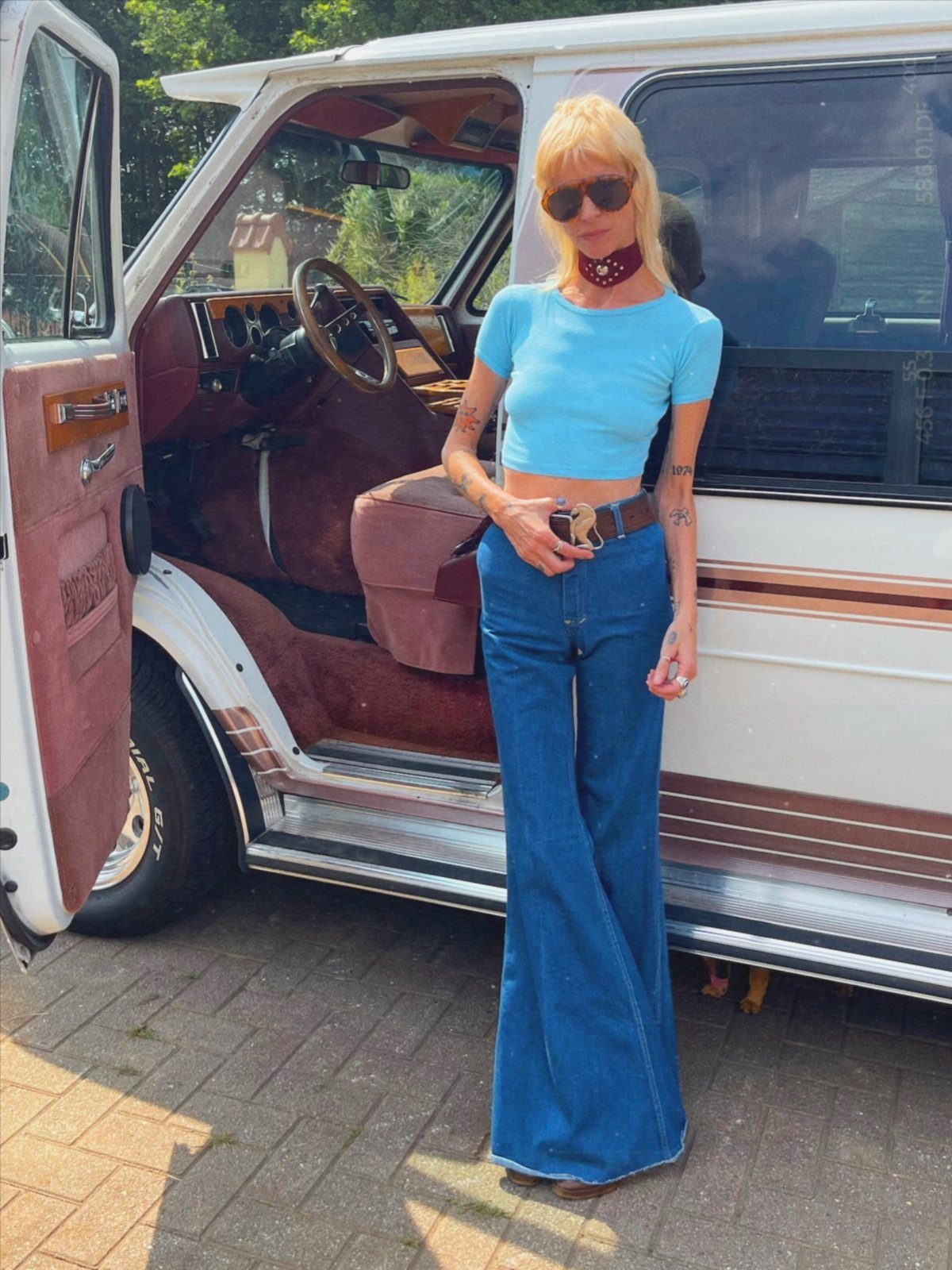 A blonde woman in a blue crop top and denim bell-bottom jeans stands by a vintage van.