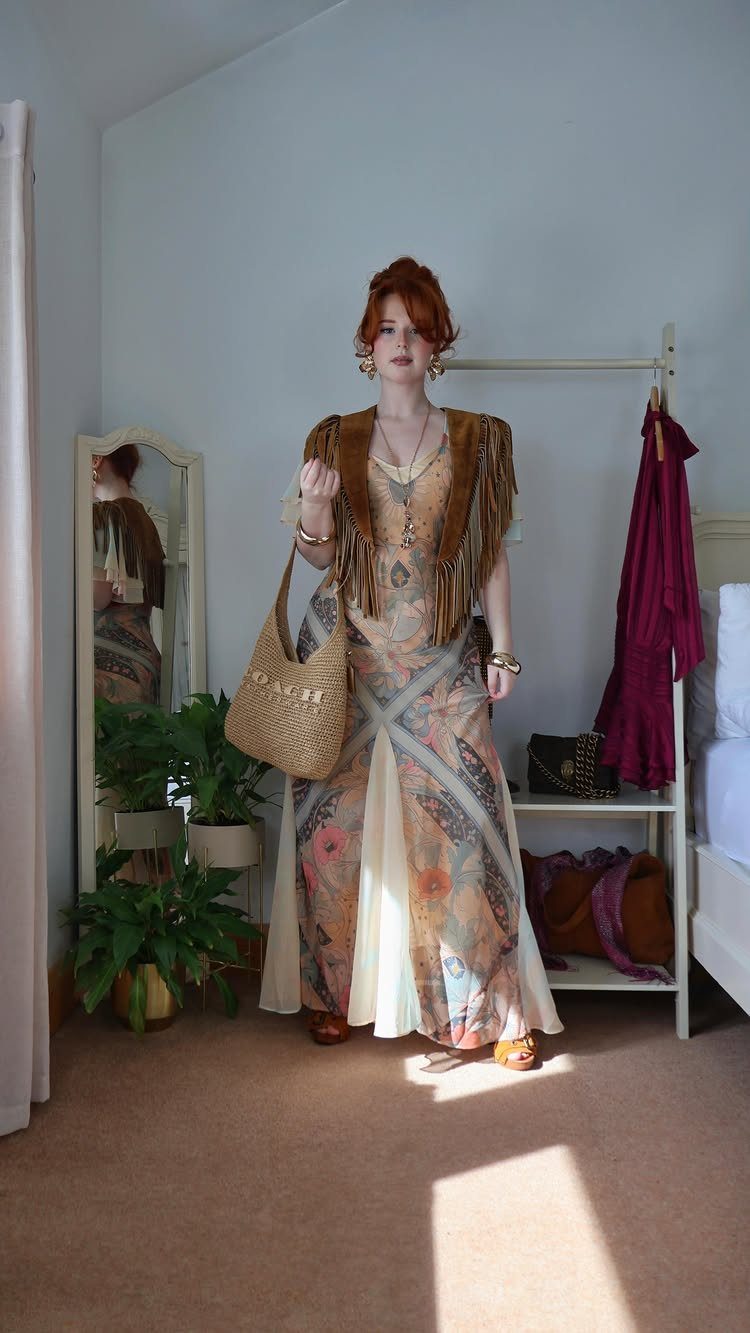 A woman wearing a long patterned bohemian dress and a suede fringe vest standing in a room.