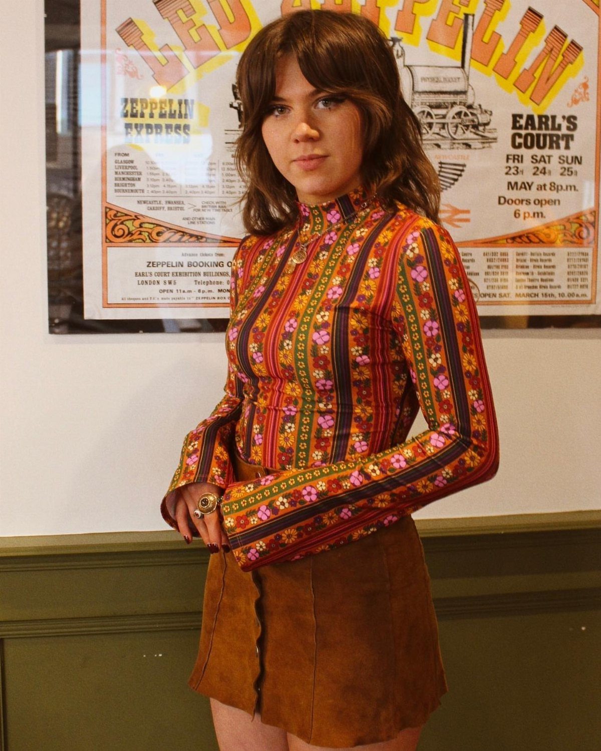 A woman wearing a colorful patterned blouse and a brown suede skirt standing before a poster.