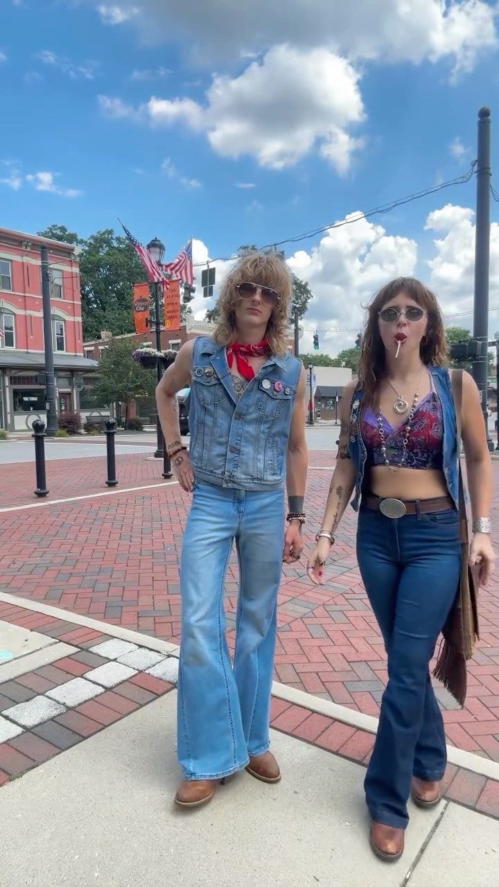 A man and woman wearing denim vests and flared jeans standing on a brick sidewalk.