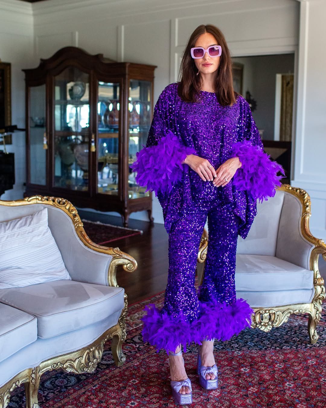 Woman in a purple sequined outfit with feather trim and pink sunglasses standing in a living room.