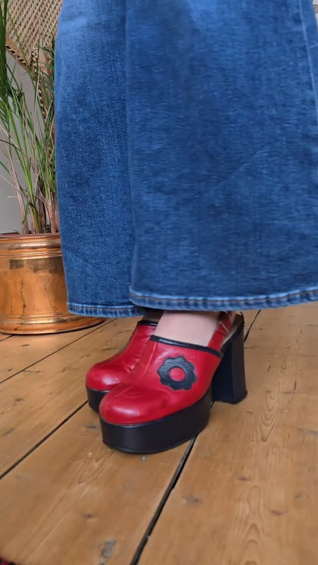 Red platform clogs with black chunky heels worn with blue denim jeans on wooden flooring.