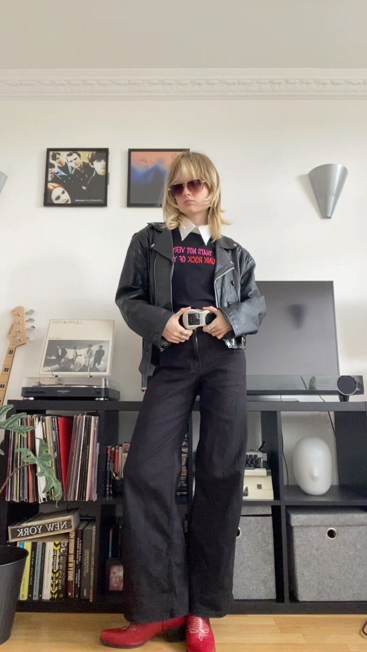 A person wearing a black leather jacket, graphic tee, and wide black trousers stands indoors.