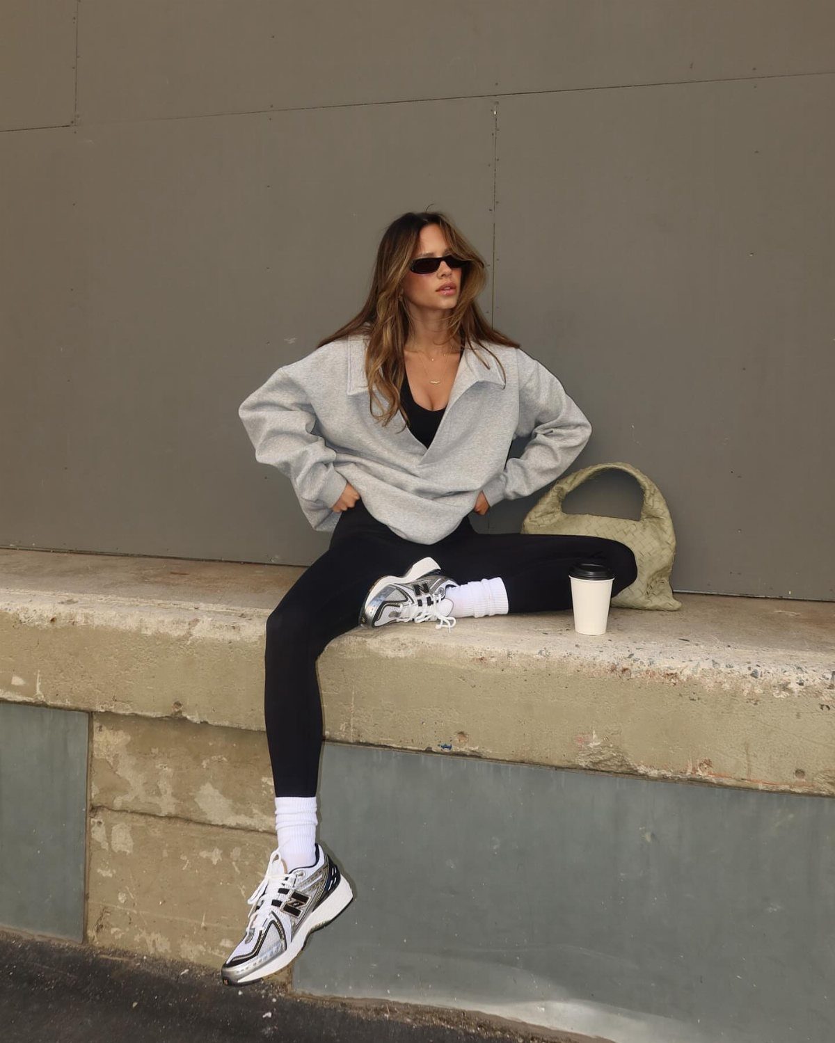 Woman in grey sweatshirt, black leggings, and chunky sneakers sitting on a concrete ledge.
