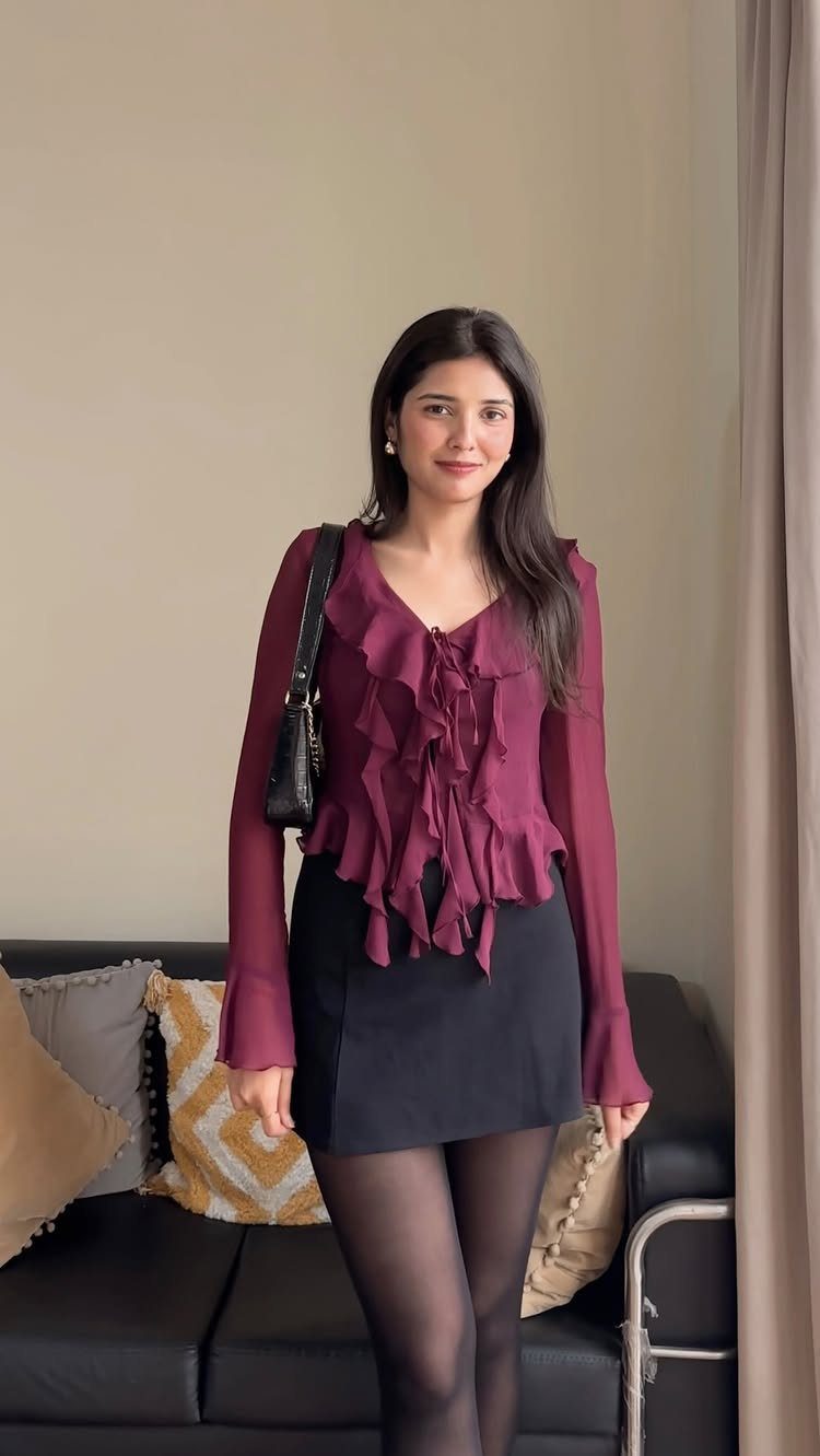 A woman wearing a burgundy ruffled blouse and a black mini skirt standing indoors.