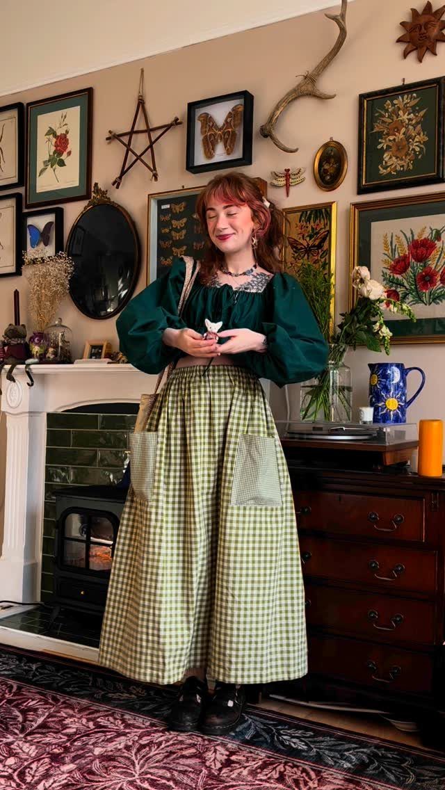 Woman in a green puff-sleeve blouse and gingham skirt standing in a vintage-style room.