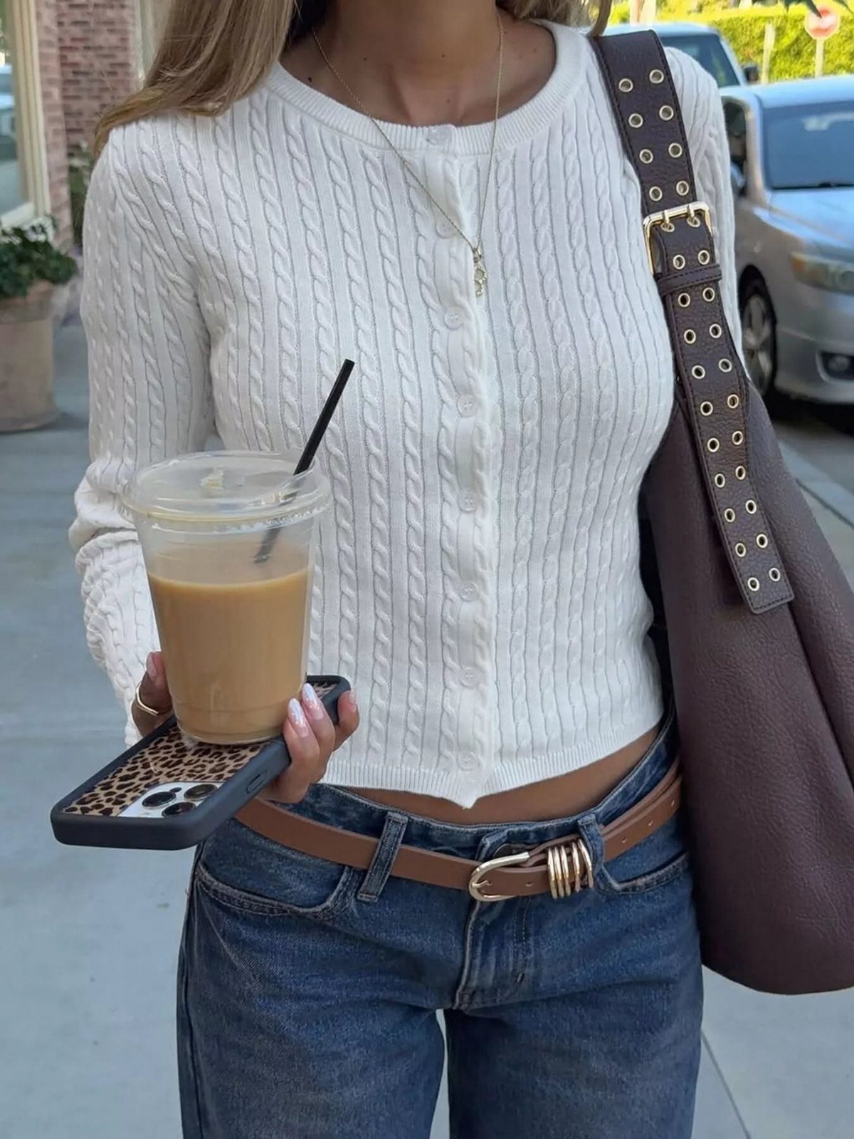 A person wearing a white cable-knit cropped top and blue jeans while holding iced coffee.
