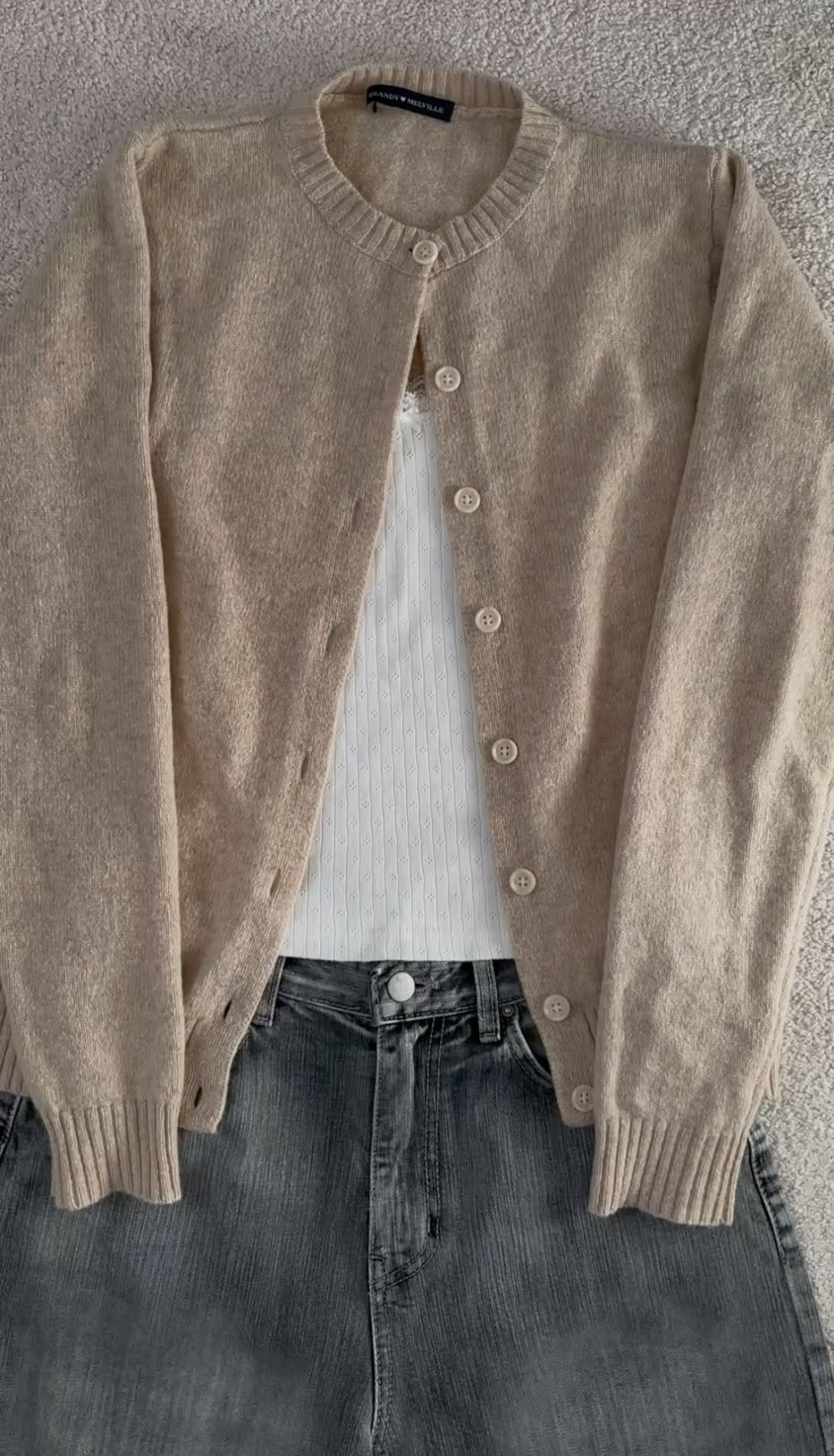 A beige knit cardigan layered over a white tank top and dark grey denim jeans.