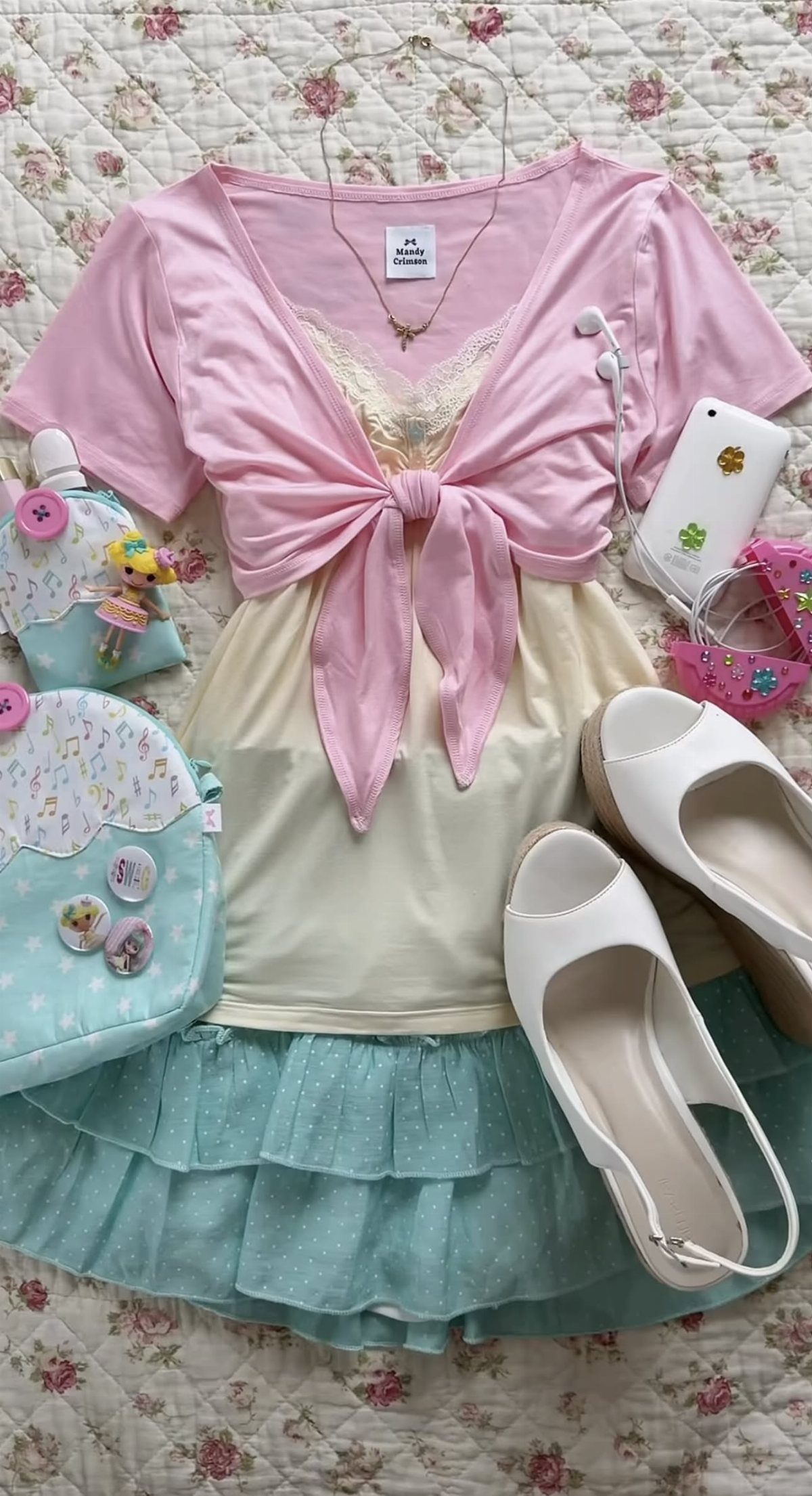 Pastel clothing items including a pink top, cream tank, and blue skirt laid out on a floral quilt.