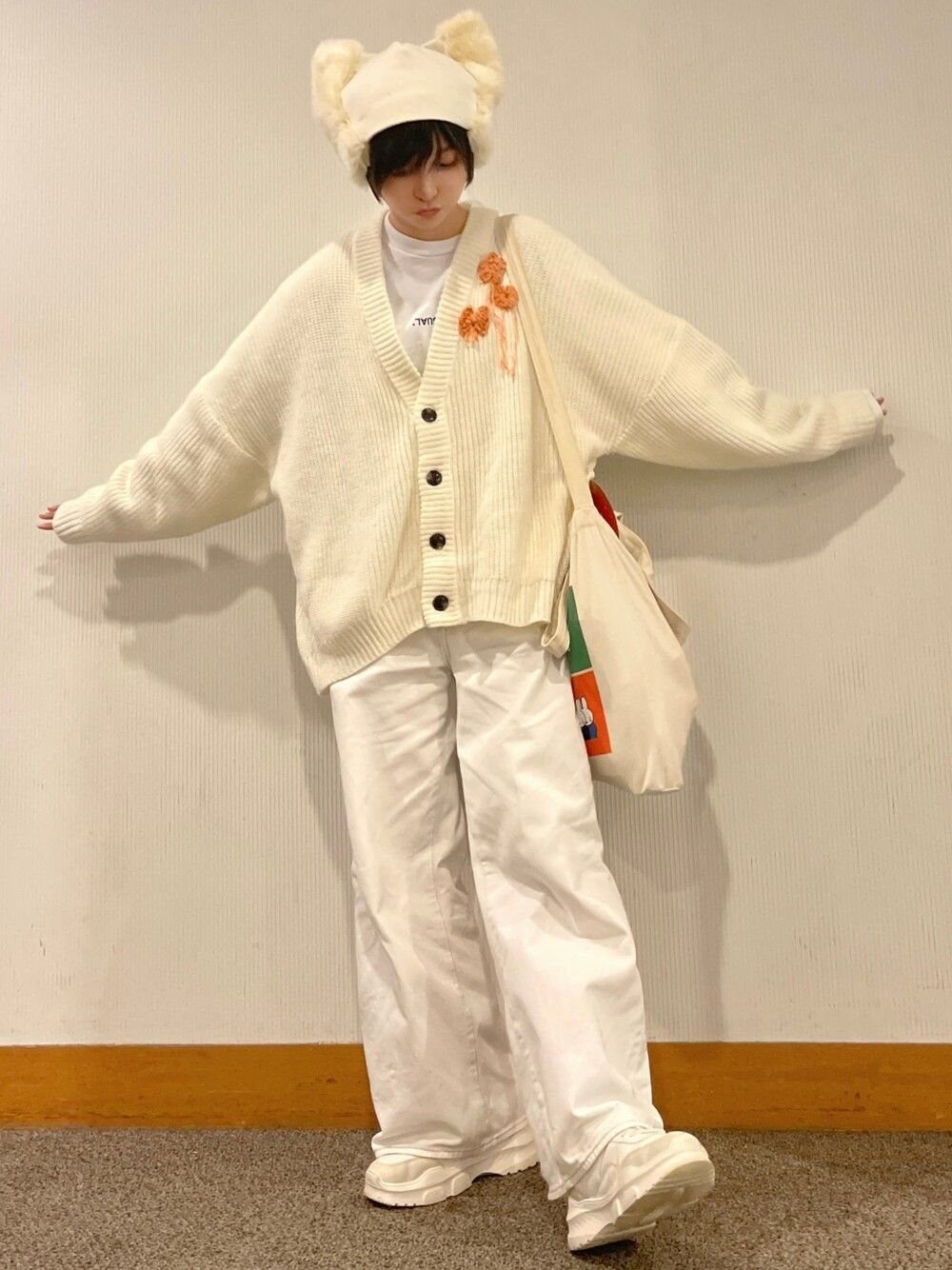 Person in oversized cream cardigan, white baggy pants, and a fuzzy bear hat standing against a wall.