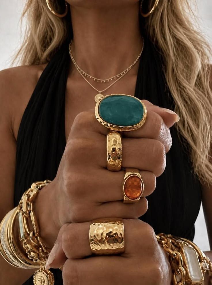 Hands wearing large gold rings with turquoise and amber stones and chunky gold bracelets.