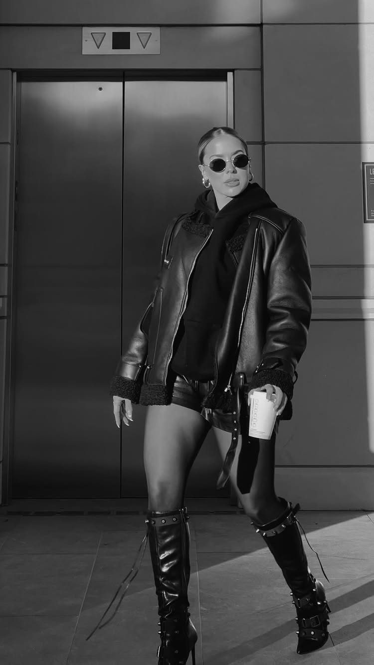 Woman wearing an oversized leather jacket, dark hoodie, shorts, and tall lace-up boots posing outdoors.