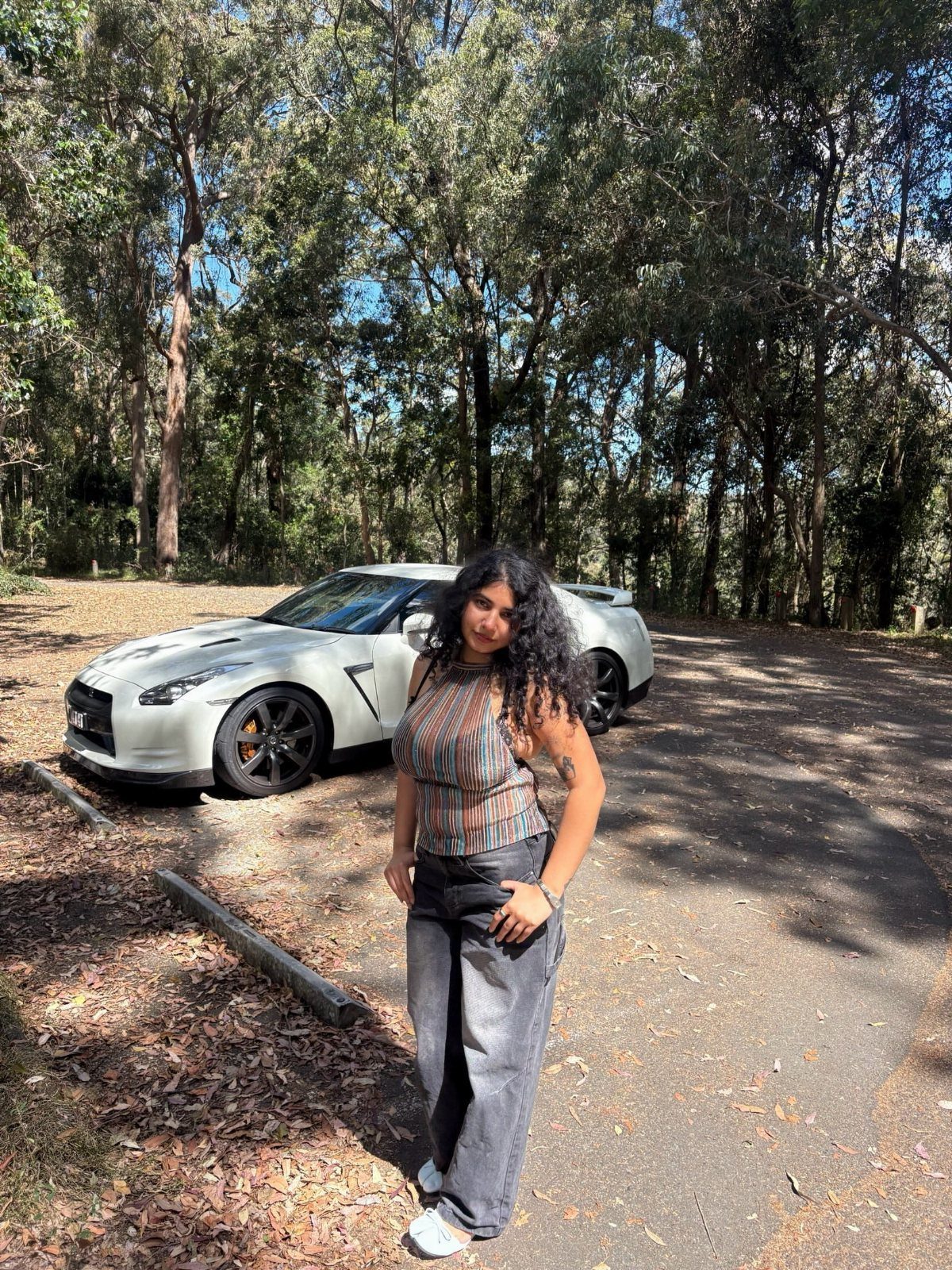 A woman in a striped top and grey trousers posing by a white sports car.
