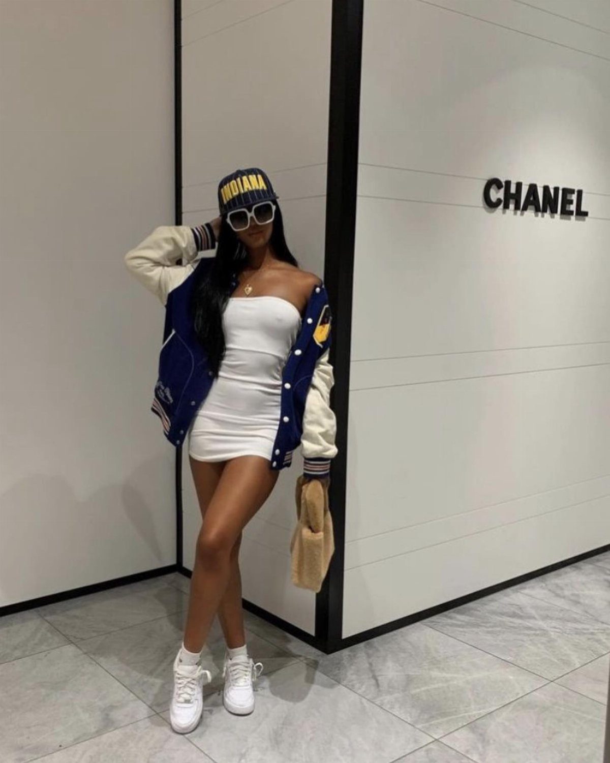 Woman wearing a white bodycon dress, varsity jacket, and white sneakers posing indoors.