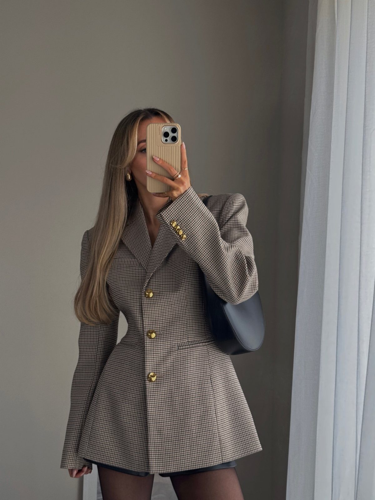 A woman posing in a mirror wearing a checkered blazer and black handbag.