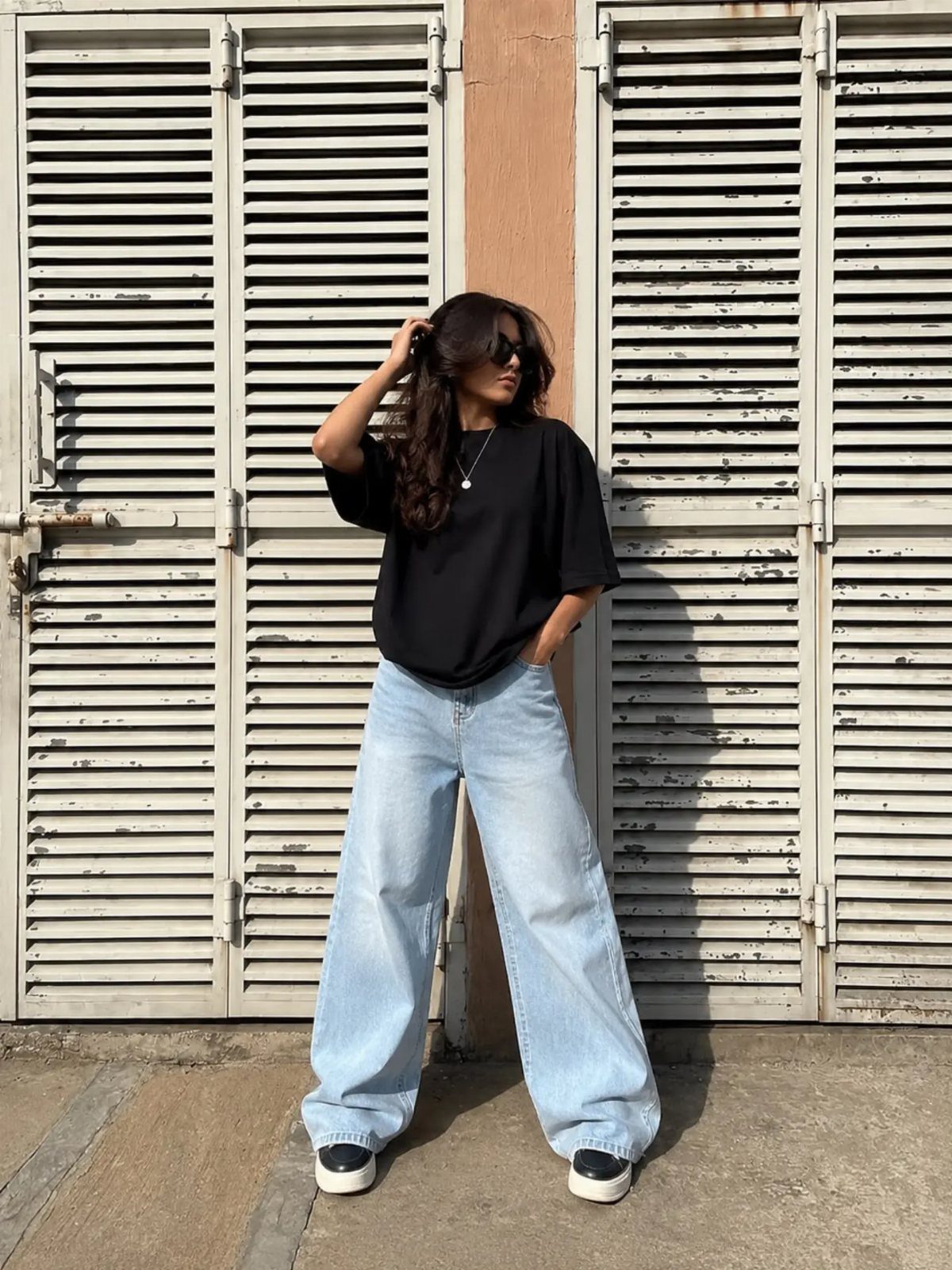 Woman posing in a black t-shirt and light wash baggy jeans against louvered doors.