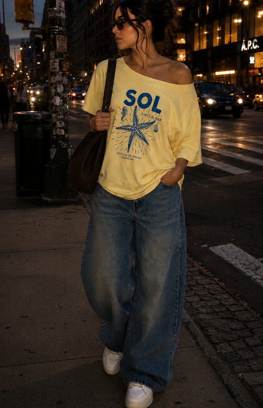 Woman in a yellow graphic t-shirt and baggy blue jeans walking on a city street at night.