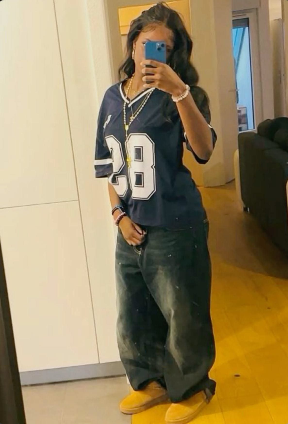 A person posing in a mirror wearing a blue jersey, baggy jeans, and tan boots.