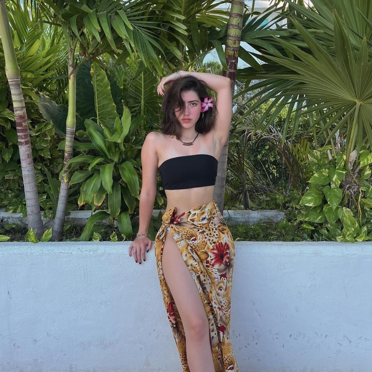 Young woman in a black bandeau top and yellow floral sarong posing against tropical plants.