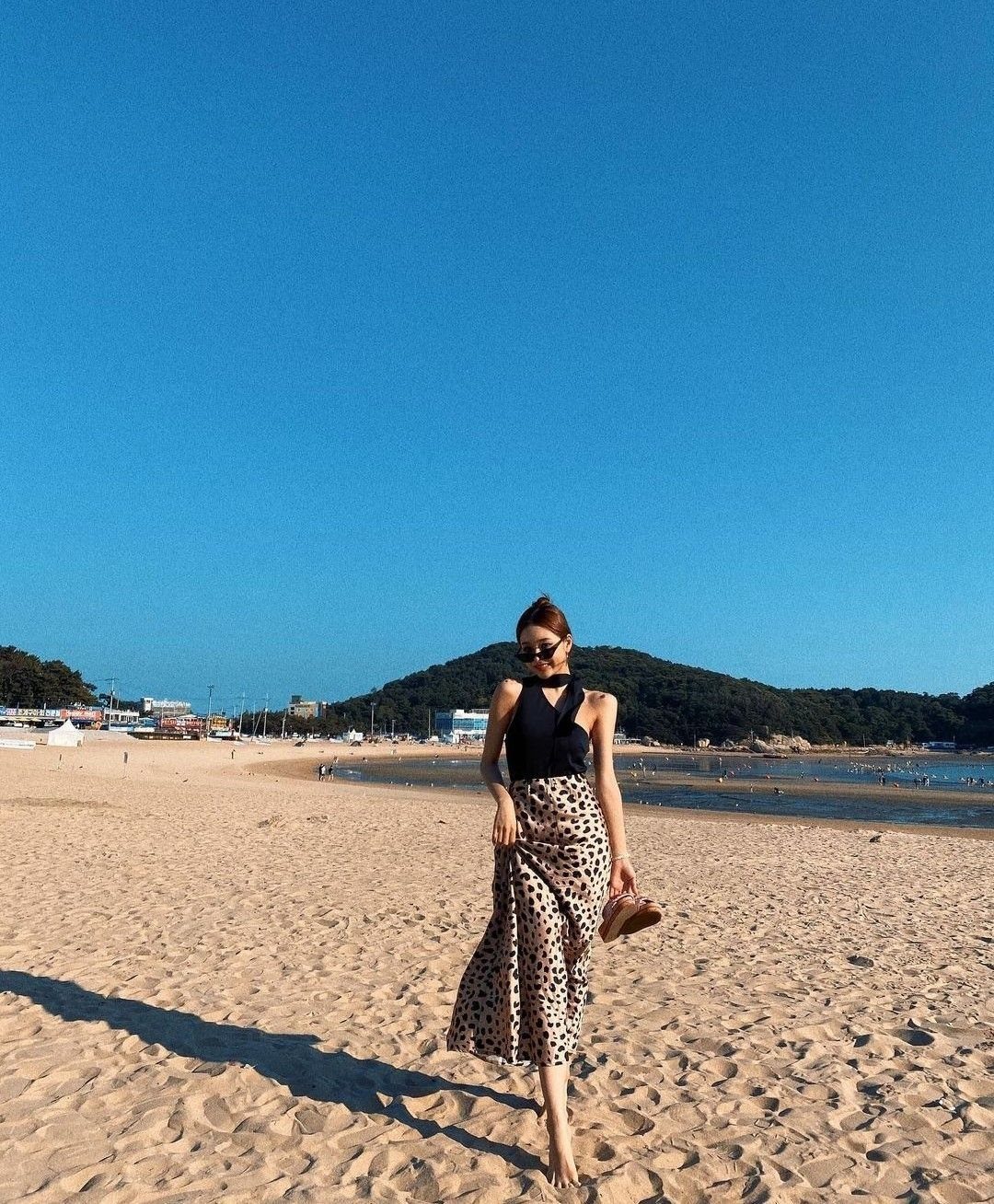 Woman in a black halter neck top and patterned skirt standing on a sunny beach.