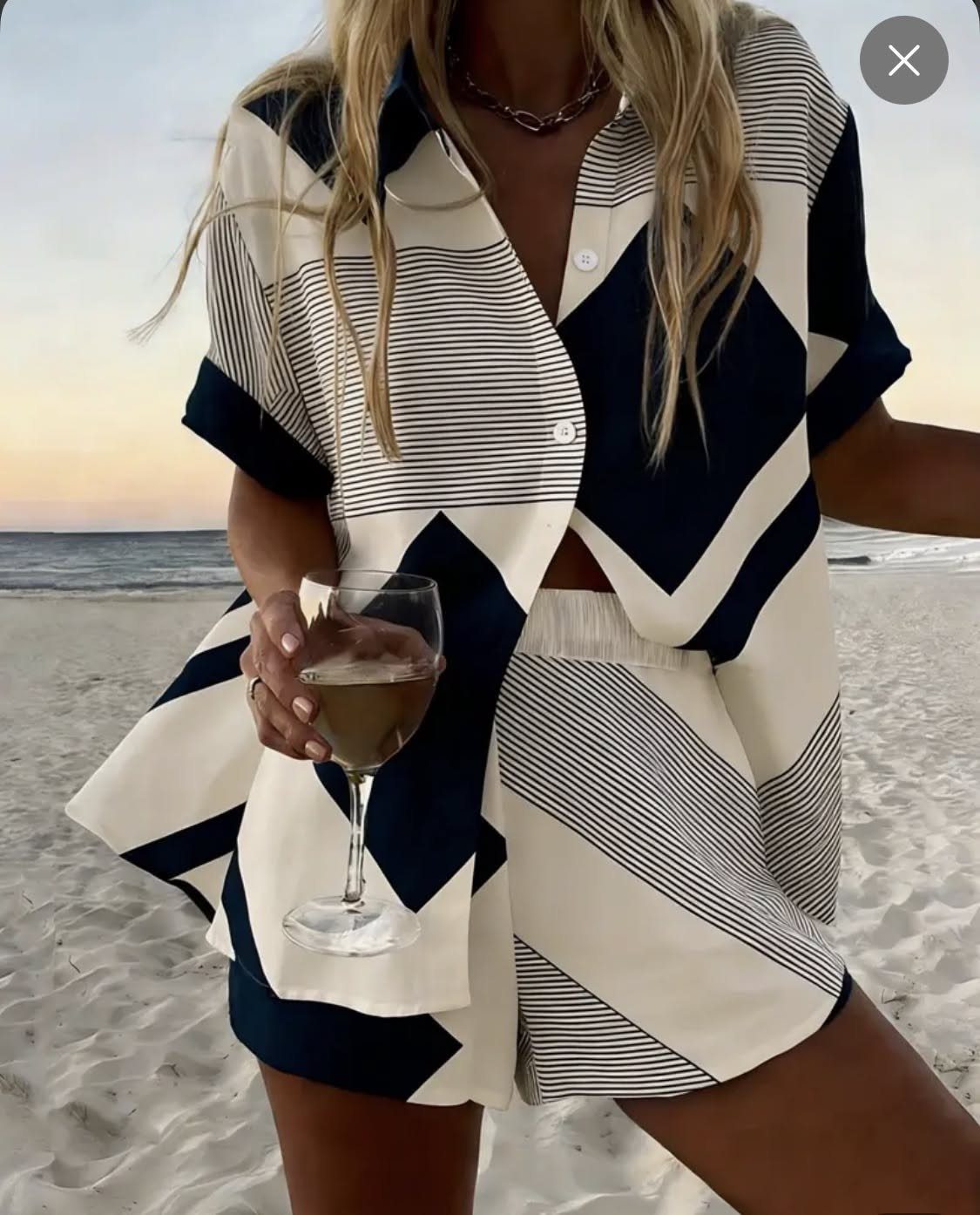 Person wearing black and white geometric patterned clothing while holding a glass on a beach.