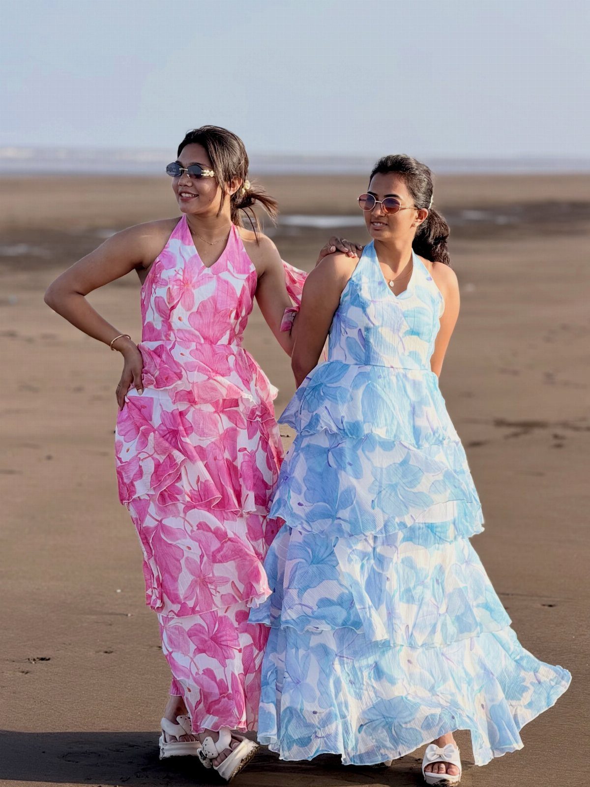 Two women in floral halter maxi dresses posing on a sandy beach.