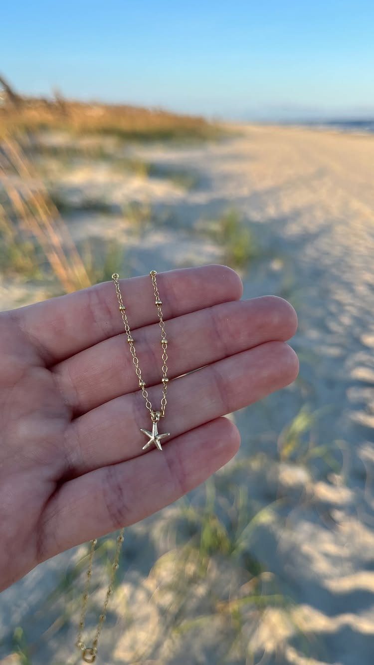 A hand holding a gold necklace with a small starfish pendant against a beach background.