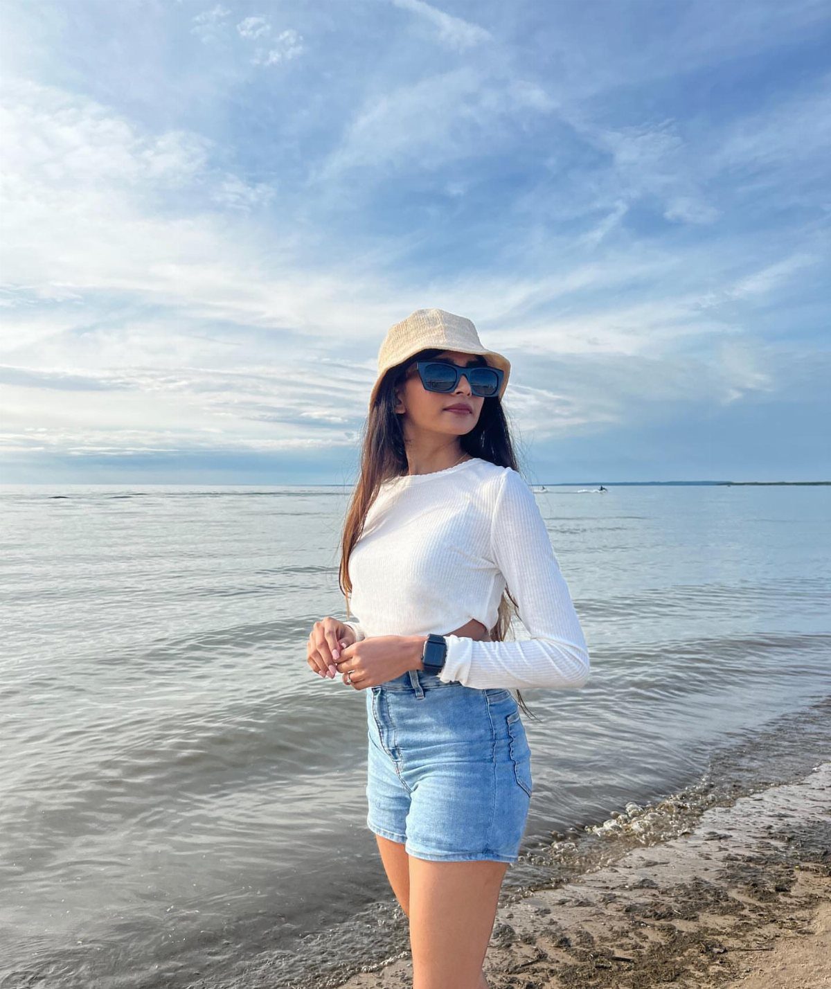 Woman in light blue denim shorts and white crop top standing on a beach.