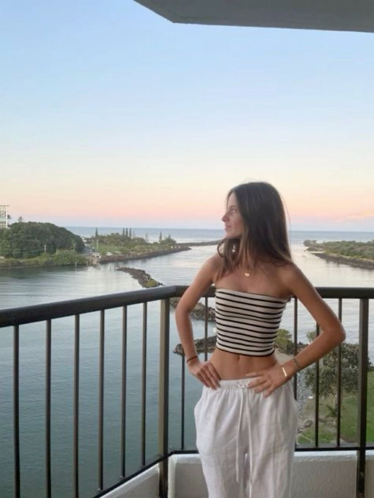 Woman in a navy striped tube top and white pants standing on a balcony overlooking the ocean.