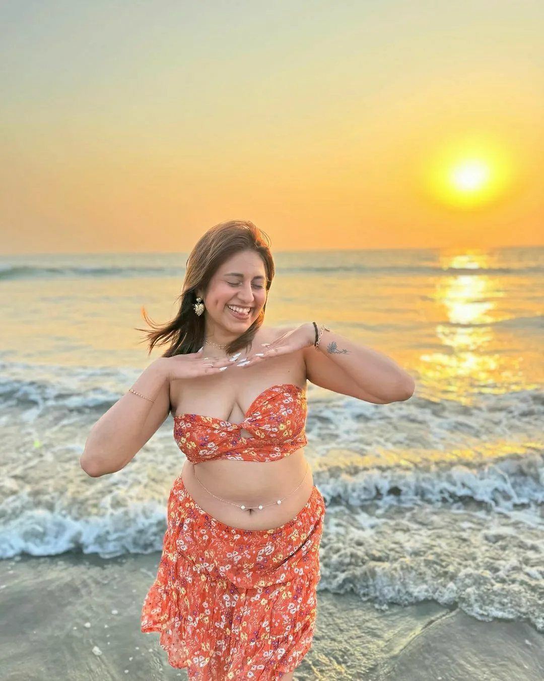 Smiling woman wearing an orange floral bikini and matching skirt on a beach at sunset.