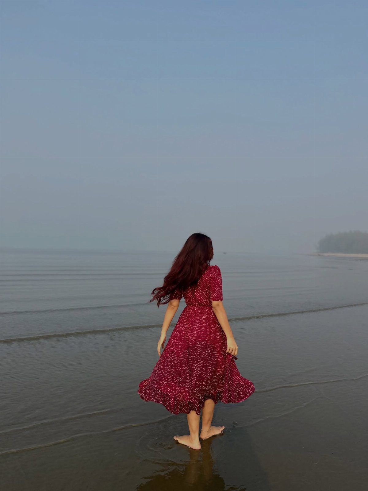 Woman in a red patterned midi dress standing in shallow ocean water at the beach.
