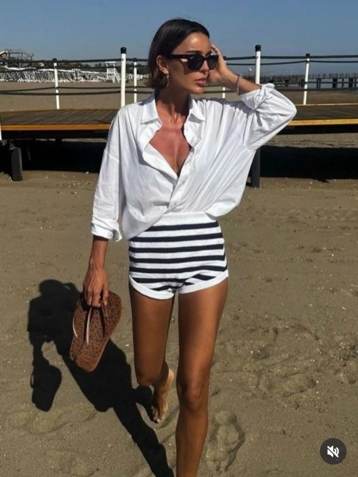 A woman in a white button-down shirt and striped shorts standing on a sandy beach.