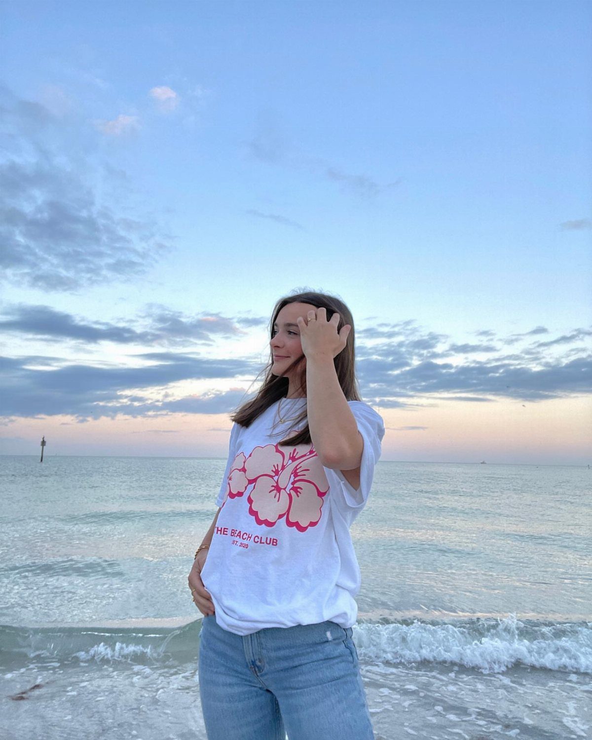 Woman wearing a white graphic t-shirt with a pink hibiscus flower on a beach.