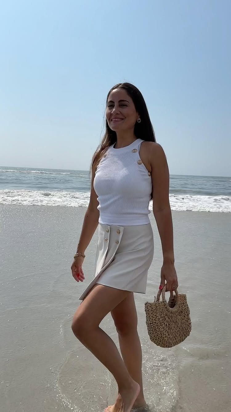 A smiling woman walking on a sunny beach wearing a white halter top and cream skirt.