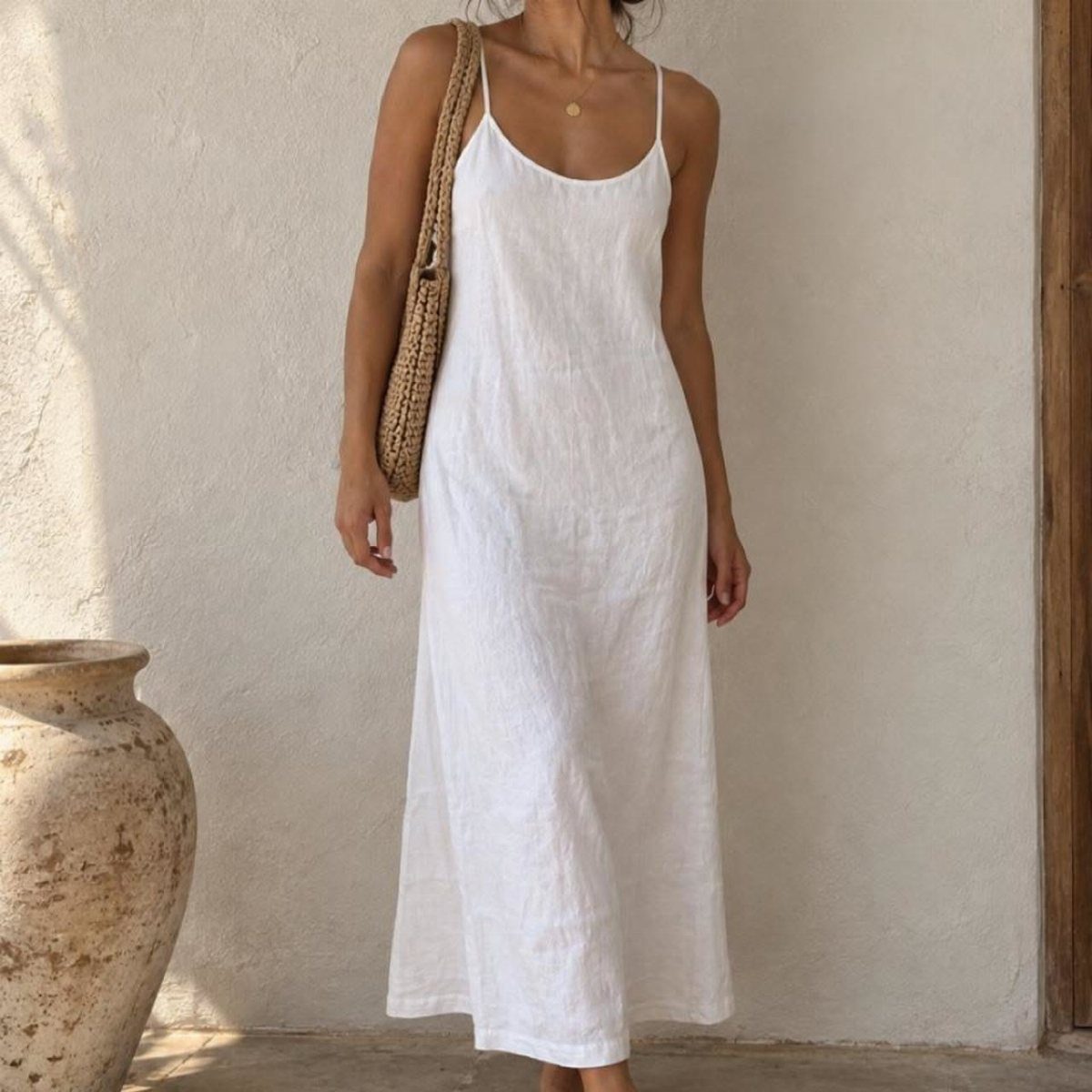 A woman wearing a white linen midi sundress and carrying a straw tote bag.