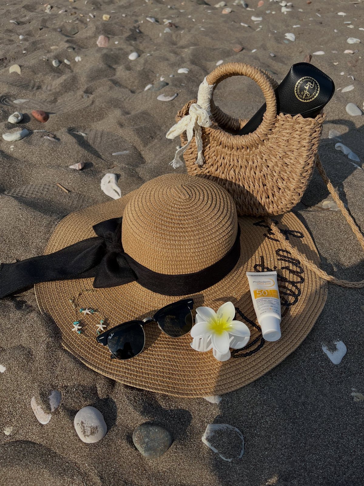 A wide-brimmed straw sun hat with sunglasses and a small woven bag on sand.