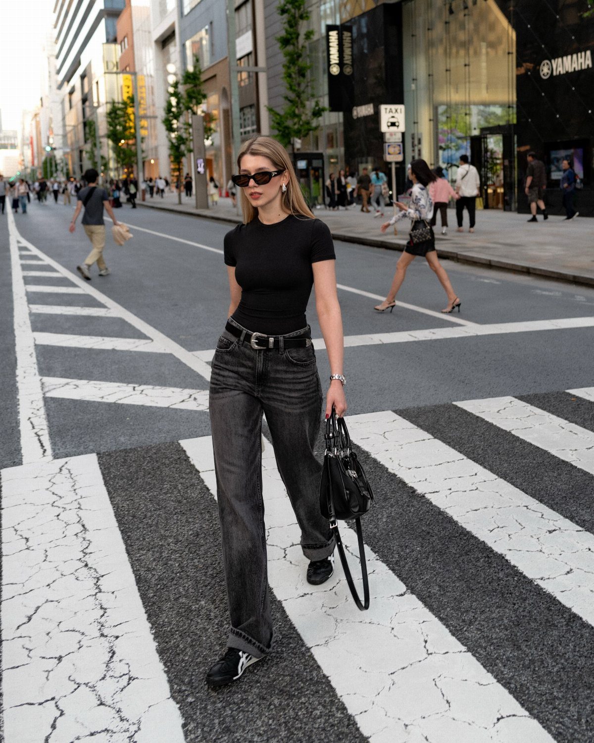 A woman in a black t-shirt and black jeans walking across a city street crosswalk.