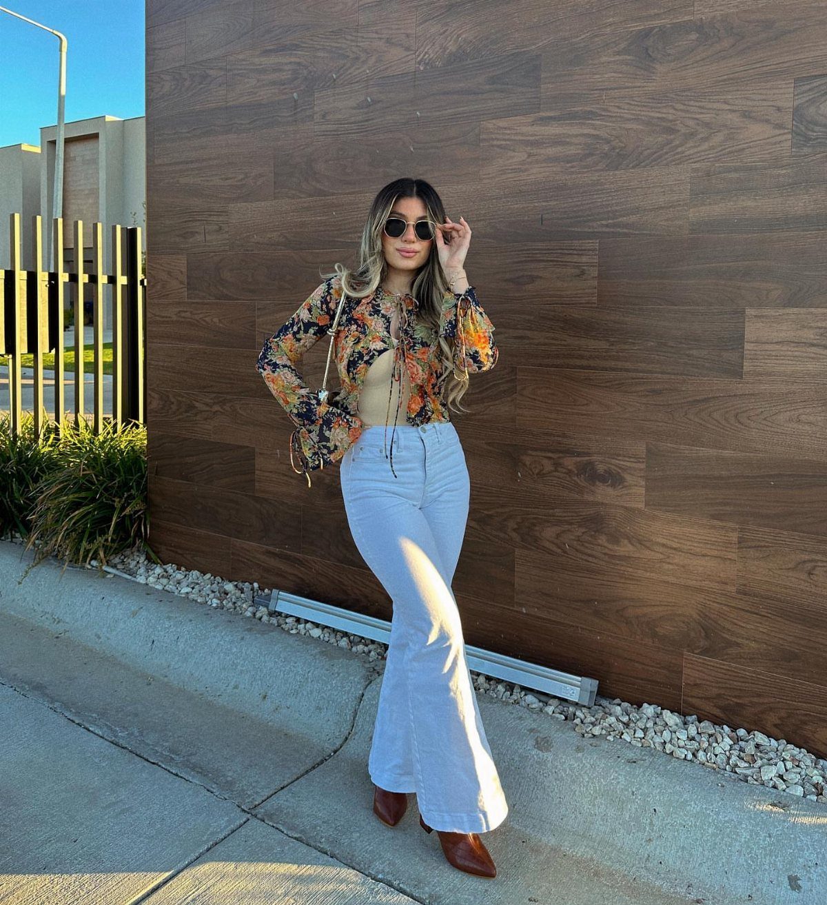 Woman in a floral blouse and white flared trousers posing against a wooden wall.