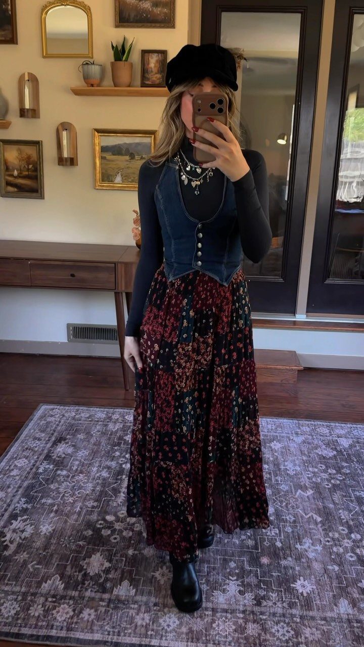 A woman posing in a mirror wearing a denim vest, floral maxi skirt, and black hat.