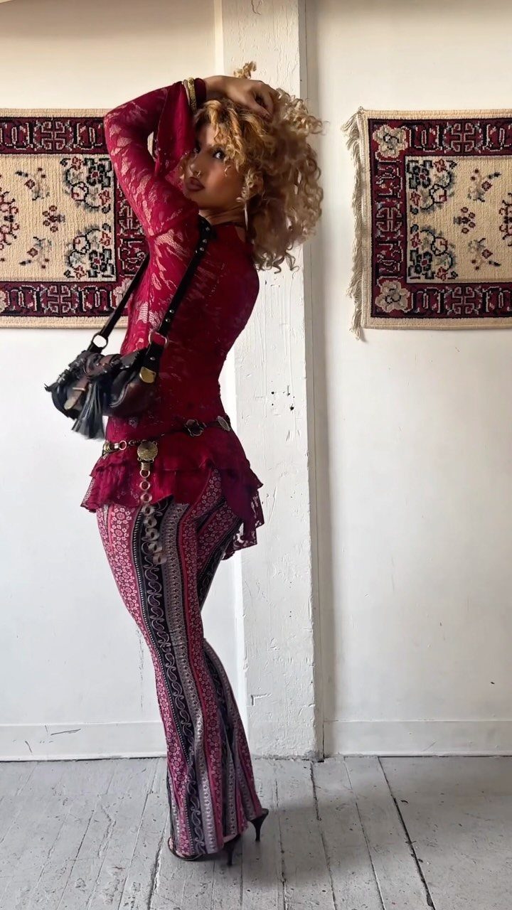 A woman in a red lace top and patterned flared trousers posing against a white wall.