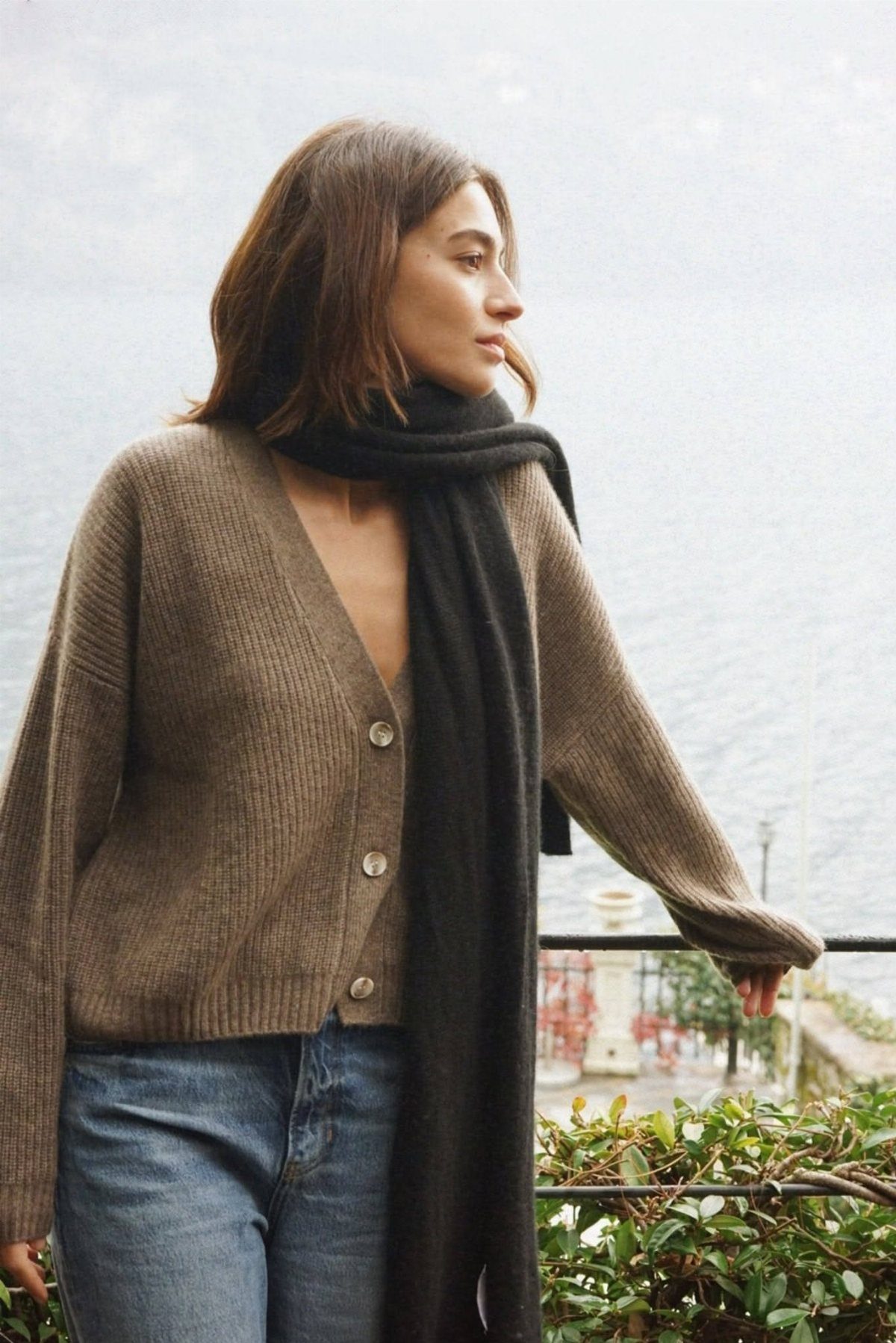 A woman wearing a brown cardigan and a long black scarf standing outdoors near water.