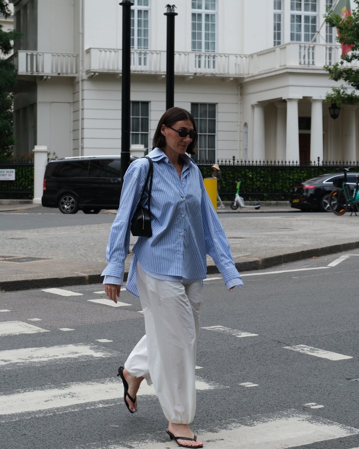 Woman walking across a street wearing a blue striped shirt, white trousers, and black high-heeled sandals.