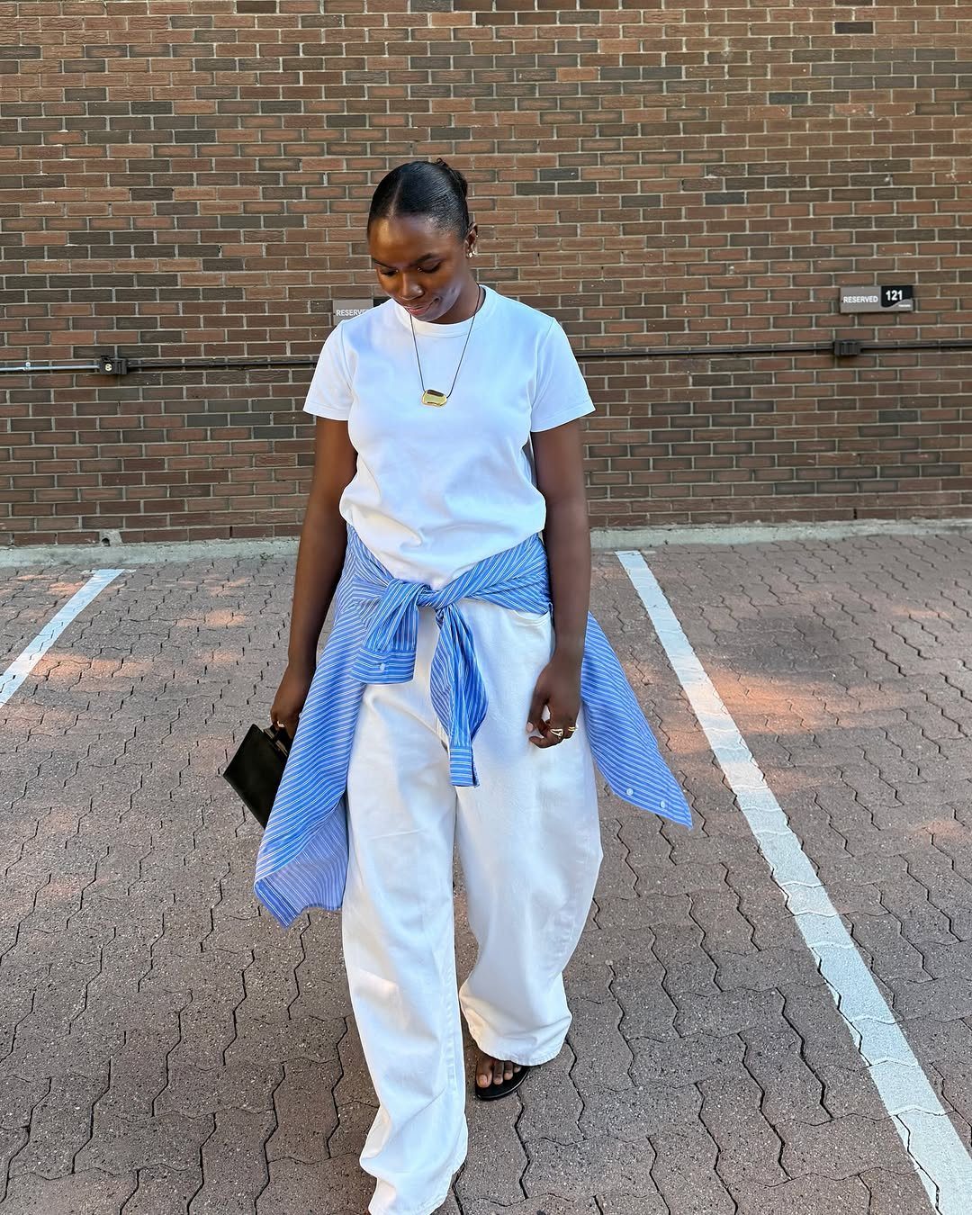 Woman in white t-shirt and wide-leg white trousers walking outdoors near a brick wall.