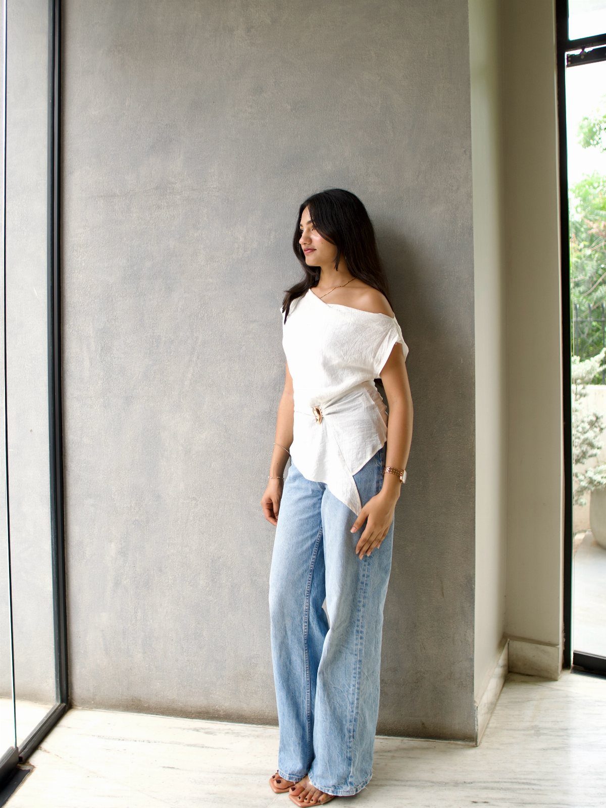 Woman in a white off-the-shoulder top and wide-leg blue jeans leaning against a grey wall.